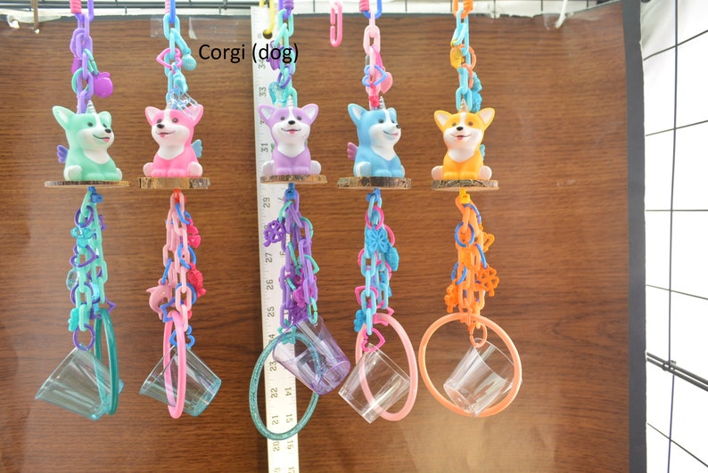 May include: Colorful hanging pet toys featuring corgi dog figurines with unicorn horns and wings. Each toy has a clear plastic cup and a ring, connected by chains in various colors like teal, pink, purple, and orange.