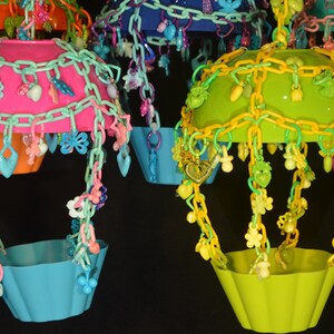 May include: Four colorful hanging bird toys with different colored plastic bowls and chains with various plastic charms. The bowls are pink, green, blue, and orange.
