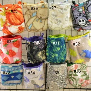 Op de afbeelding: Een rasterweergave van kleine fleece zakjes met verschillende patronen. Ontwerpen omvatten pompoenen, teddyberen, schedels en stripfiguren. Andere zakjes hebben aardbeien, bloemen en geometrische patronen. Elk zakje heeft een andere kleurafwerking.