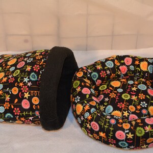 Puede incluir: Dos camas para mascotas. Una es una cama cerrada, negra con interior negro. La otra es una cama redonda y abierta. Ambas están cubiertas con un estampado floral con flores naranjas, rosas, azules y amarillas.