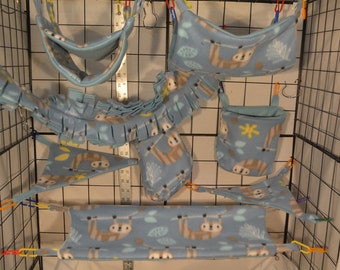 Puede incluir: Un conjunto de accesorios para mascotas de forro polar azul, que incluye hamacas, una bolsa y un puente con flecos. Los artículos presentan un patrón repetido de perezosos y hojas. Los accesorios están diseñados para colgarse dentro de una jaula.