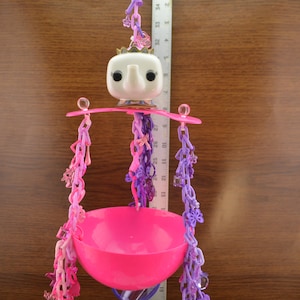 May include: A pink bird toy with a white elephant figurine on top. The toy has multiple purple and pink chains with charms hanging from it. The toy is hanging from a ceiling.