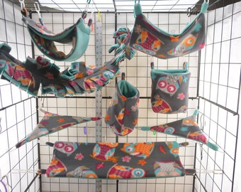 Puede incluir: Un conjunto de nueve accesorios para jaulas de mascotas colgantes con temática de búhos. Los accesorios están hechos de tela suave y presentan una variedad de colores, incluyendo gris, azul verdoso, naranja, rosa y blanco. Los accesorios incluyen una hamaca, un túnel, una bolsa y varios juguetes colgantes.