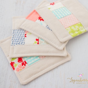 May include: Four square fabric coasters with a patchwork design. Each coaster has sections of floral and polka dot patterns in red, blue, pink, and green, set against a neutral background. The edges are finished with a beige border.