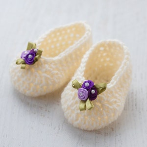 Könnte beinhalten: Ein Paar weiße gehäkelte Babyschuhe mit violetten und weißen Blumenverzierungen.