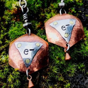 May include: A pair of copper and silver earrings with a hammered finish. Each earring features a triangular shape with the letter "e" and a dash inscribed on it. The earrings are suspended from silver hooks.