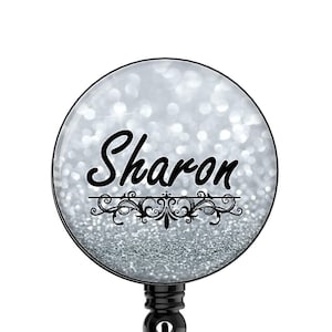 May include: A silver glitter retractable badge holder with a black border and the name "Sharon" printed in black cursive font.