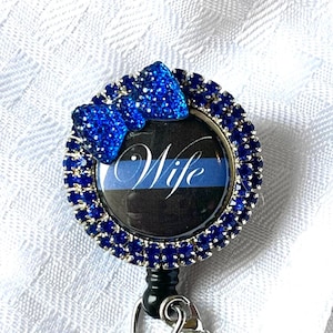 May include: A blue badge reel with a blue bow and a circle of blue rhinestones. The badge reel has the word "Wife" on it. There is a blue heart and a silver charm with handcuffs and a heart attached to the reel.