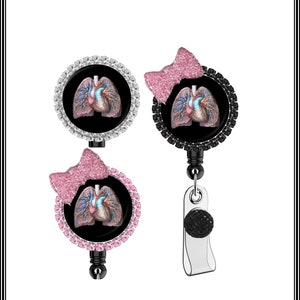 May include: A black and white anatomical heart and lungs image on a black background with a pink glitter bow on top. The image is surrounded by rhinestones and is attached to a retractable badge holder with a black rhinestone clip.