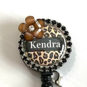 May include: A black and brown leopard print retractable badge holder with a black flower charm, a black bead, and a brown bead. The badge holder has a silver metal ring and a clear plastic strap. The badge holder says "Kendra" on it.