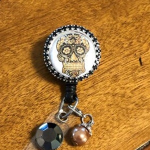 May include: A black and silver retractable badge holder with a round, black and white skull design. The skull has gold details and floral designs. The badge holder has a black crystal and a brown pearl charm.