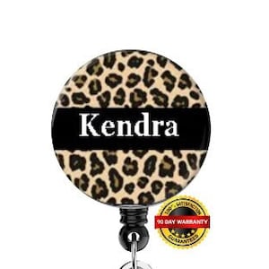 Puede incluir: Un portagafete retráctil con un diseño de estampado de leopardo y el nombre "Kendra" impreso en blanco sobre un fondo negro. El portagafete tiene un clip de plástico transparente y un pivote de metal negro.