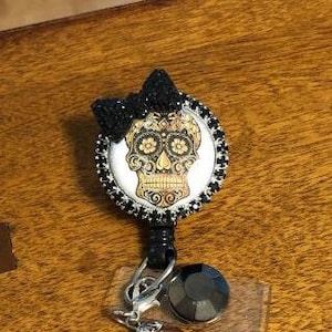 May include: A black and white sugar skull with gold accents and a black rhinestone bow. The skull is surrounded by black rhinestones and is attached to a retractable badge holder with a black rhinestone charm and a pearl charm.