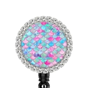 May include: A retractable badge reel with a round, iridescent mermaid scale design. The design is surrounded by a silver rhinestone border. A small, purple pearl charm hangs from the reel, and the clip has a pink glitter accent.