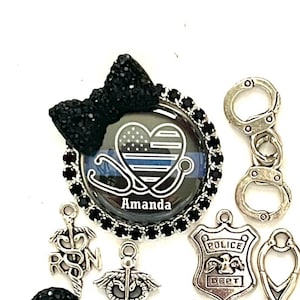 以下が含まれることがあります： 黒いリボン、ハート型の星条旗デザインのバッジ、手錠、医療シンボルなど、チャームとアクセサリーのコレクション。バッジは「Amanda」という名前でパーソナライズされています。「Pick 2 charms」というテキストも表示されています。