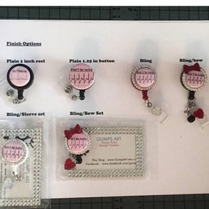 Puede incluir: Una variedad de opciones de carretes de insignia con diferentes acabados. Los carretes presentan un dise&ntilde;o rosa y blanco con el texto "Don't be tachy" y un gr&aacute;fico de ritmo card&iacute;aco. Los acabados incluyen carrete simple de 2,5 cm, bot&oacute;n simple de 3,175 cm, bling, bling/lazo y bling/manga.