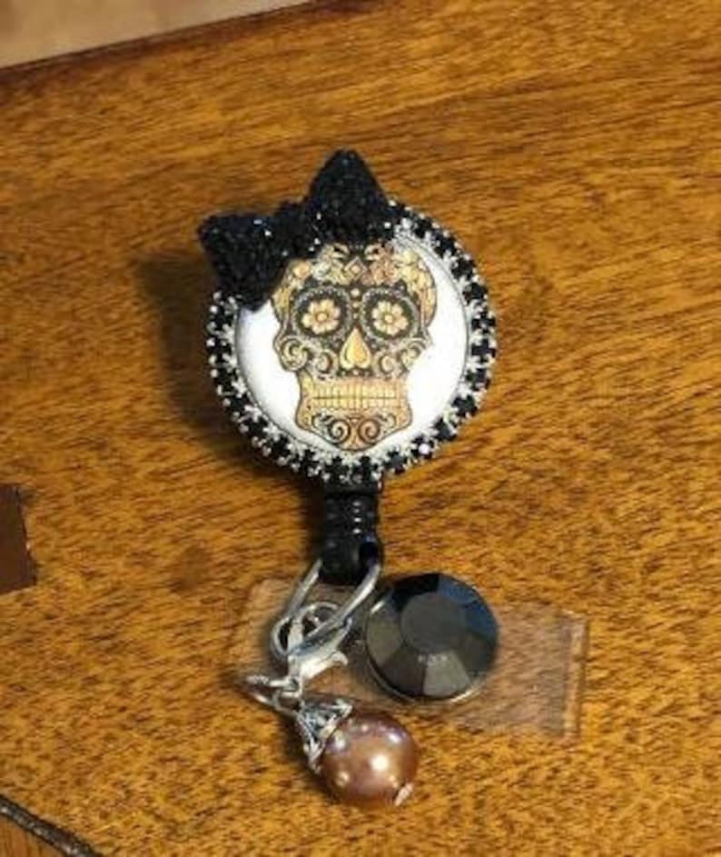 May include: A black and white sugar skull with gold accents and a black rhinestone bow. The skull is surrounded by black rhinestones and is attached to a retractable badge holder with a black rhinestone charm and a pearl charm.