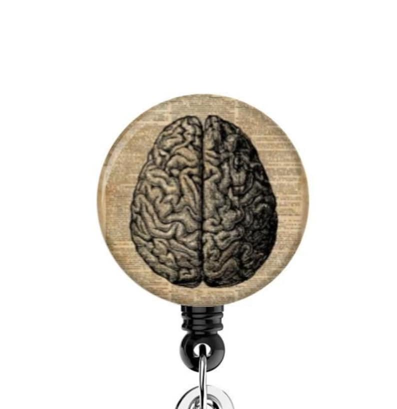 May include: A round, vintage-style badge reel featuring a detailed illustration of a human brain. The brain image is in black and white, set against a background of aged paper. The reel has a black retractable cord and a metal clip.