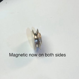 Pode incluir: Um bot&atilde;o magn&eacute;tico prateado com uma base de pl&aacute;stico branco. O texto "Magnetic now on both sides" &eacute; vis&iacute;vel abaixo do bot&atilde;o.