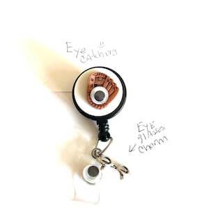 May include: A baseball glove design on a retractable badge reel. The reel has a white face with a black border. A small charm of eyeglasses is attached. The text "Eye Catching" and "Eye Glasses Charm" are handwritten.