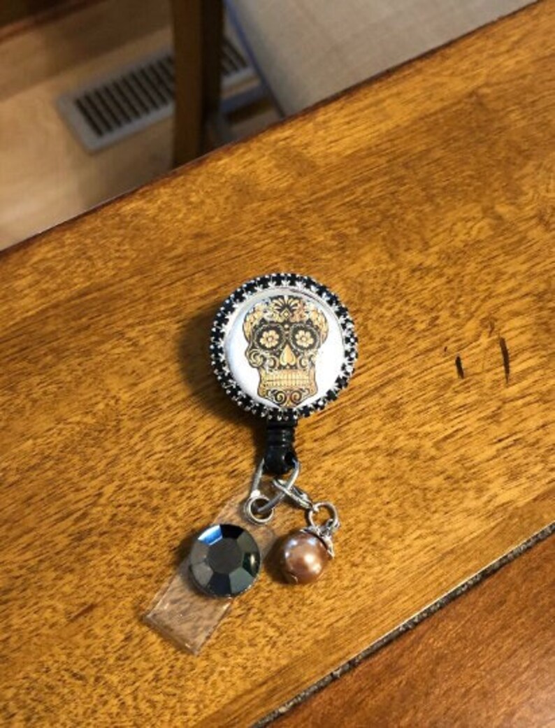 May include: A black and silver retractable badge holder with a round, black and white skull design. The skull has gold details and floral designs. The badge holder has a black crystal and a brown pearl charm.