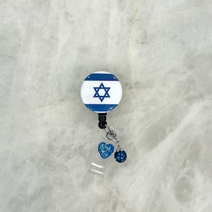 May include: A white and blue retractable badge holder with a blue Star of David on the front. The holder has a blue glitter heart and a dark blue pearl charm attached.