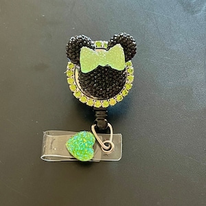 May include: A black and green rhinestone badge reel in the shape of a mouse head with a green bow. The badge reel has a clear plastic strap with a green heart-shaped accent.