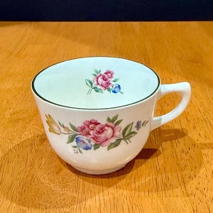 May include: A white ceramic teacup featuring a delicate floral pattern with pink roses, blue flowers, and green leaves. The rim of the cup is accented with a thin green line, and a smaller floral motif is visible on the interior wall of the cup.
