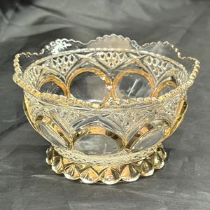 May include: A clear glass bowl with gold accents. The bowl has a scalloped rim and a decorative pattern. The gold accents are on the rim and around the base of the bowl. The bowl is sitting on a dark surface.