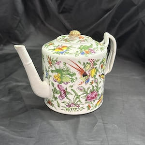 May include: White porcelain teapot with a floral and bird design. The teapot features a curved spout, a handle, and a lid with a gold-colored knob. The design includes colorful flowers, birds, and foliage, creating a decorative and elegant appearance.