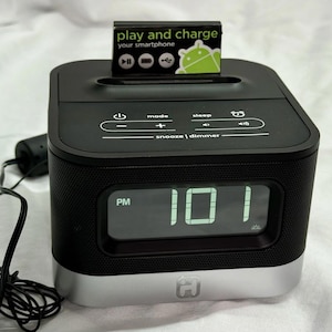 以下が含まれることがあります： 黒とシルバーのデジタル目覚まし時計。スマートフォン充電ドックが内蔵されています。 時計は、大きな緑色のデジタル数字で時刻を表示します。 時計の上部には「play and charge your smartphone」という文字があります。