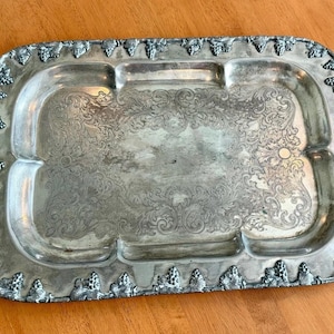 May include: Silver-toned rectangular serving tray with ornate floral and scrollwork detailing. The tray has two handles and a decorative border with grape cluster accents. The tray is approximately 18 inches long and 12 inches wide.