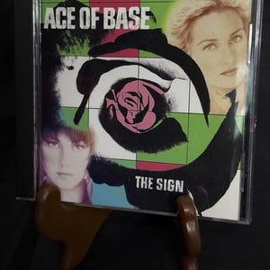 May include: A CD case for the album "The Sign" by Ace of Base. The cover features the band's name, a stylized rose, and images of the band members. The background is a mix of green, pink, and black.