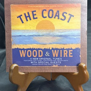 May include: A CD case with a sunset and ocean scene, with the text "THE COAST WOOD & WIRE" in blue and white. The case is displayed on a small wooden easel. The text also includes "11 NEW ORIGINAL TUNES".