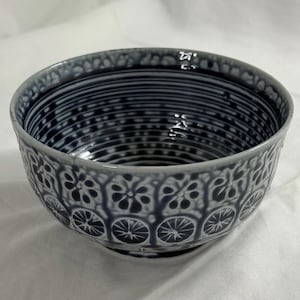 May include: A ceramic bowl with a dark blue and white pattern. The bowl has a repeating floral and geometric design around the rim and base, with horizontal lines and dots inside. The bowl is likely used for serving food.