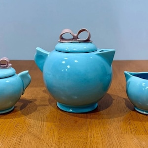 May include: A three-piece ceramic tea set in bright turquoise blue. The set includes a teapot, a lidded sugar bowl, and a creamer. Both the teapot and sugar bowl lids feature a small, sculpted light pink bow handle on top.