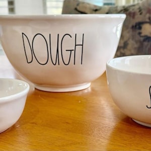 以下が含まれることがあります： 「DOUGH」、「SAUCE」、および「CHEESE」と書かれた黒い文字が特徴の、3つの白いセラミックボウル。ボウルはさまざまなサイズで、木製のテーブルの上に置かれています。背景には窓と模様のある椅子があります。