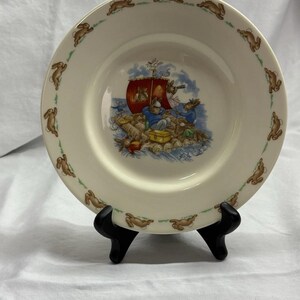May include: Cream-colored ceramic plate featuring a nautical scene with rabbits in a boat. The plate's rim is decorated with brown rabbit silhouettes. The central illustration depicts rabbits on a raft, with a red sail, sailing on blue water.
