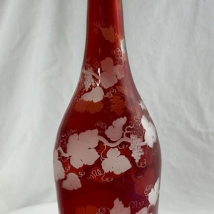 May include: A ruby red glass decanter with a matching stopper. The decanter is adorned with a white grape vine design. The stopper is rectangular. This decorative bottle is likely used for wine or spirits.