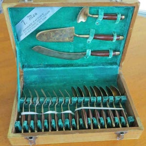 Peut inclure: Ensemble de couverts vintage dans un coffret en bois. L'ensemble comprend des ustensiles de service et des fourchettes et cuillères individuelles. Les manches sont en bois foncé et le métal est doré. L'intérieur du coffret est doublé de velours vert.