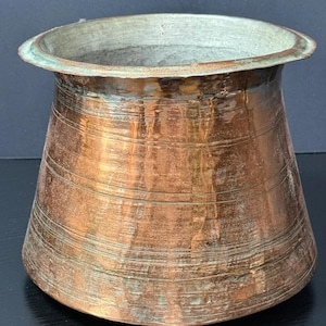 May include: A copper-colored, antique-style vase with a wide, slightly flared rim. The vase has horizontal bands and a weathered patina, suggesting age and use. The interior is a lighter, greenish-gray color.
