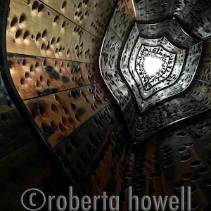 May include: A wooden spiral staircase with a light at the top. The staircase is made of wood planks with holes drilled in them. The holes are arranged in a pattern that creates a visual effect of depth and movement.