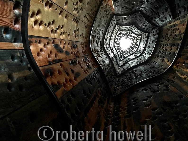 May include: A wooden spiral staircase with a light at the top. The staircase is made of wood planks with holes drilled in them. The holes are arranged in a pattern that creates a visual effect of depth and movement.