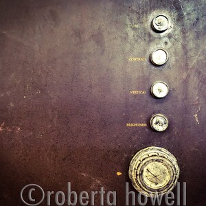 以下が含まれることがあります： 5つの丸いノブが付いたビンテージテレビのコントロールパネルのクローズアップ。ノブには「明るさ」、「垂直」、「コントラスト」と、ラベルのないノブが2つあります。パネルはダークブラウンで、表面はテクスチャ加工されています。