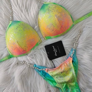 May include: A two-piece bikini set with a vibrant, iridescent color scheme of yellow, orange, and green. The bikini top and bottom are embellished with silver rhinestone accents. The set includes a tag that reads "Dana Carmont Elites".