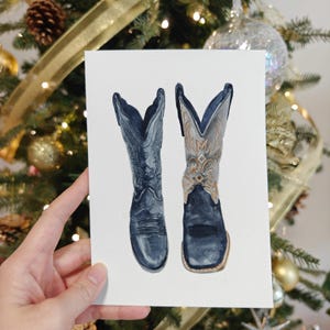 K&ouml;nnte beinhalten: Ein Aquarellbild von zwei blauen Cowboystiefeln mit goldener Stickerei. Die Stiefel befinden sich auf einem wei&szlig;en Hintergrund.