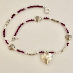 Puede incluir: Un collar de plata con un colgante de corazón martillado y tres dijes más pequeños en forma de corazón. El collar está ensartado con pequeñas cuentas redondas de color rojo oscuro y perlas blancas. Un cierre de palanca asegura el collar.