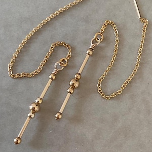 Op de afbeelding: Een paar gouden kettingoorbellen met een lange, dunne staaf en drie gouden kralen. De oorbellen zijn verbonden met een ketting die als ketting gedragen kan worden.