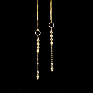 May include: A pair of gold-colored dangle earrings against a black background. Each earring features a delicate chain with a small hoop and a series of small, round beads. The earrings are designed to hang down, creating a graceful, elongated look.