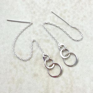 Puede incluir: Un par de pendientes de hilo plateados. Cada pendiente presenta una cadena que se enhebra a través de la oreja, con tres anillos plateados conectados colgando al final. Los pendientes se muestran sobre una superficie clara.